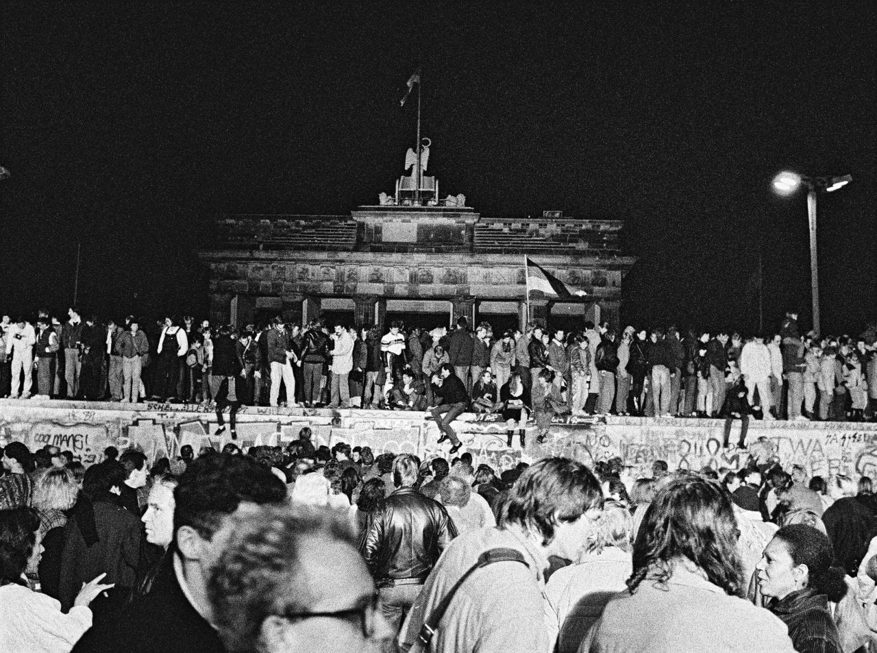 Fotos von feiernden Menschen auf und vor der Berliner Mauer bei Nacht. Zentral im Bild ist das Brandenburger Tor hinter der Mauer zu sehen.