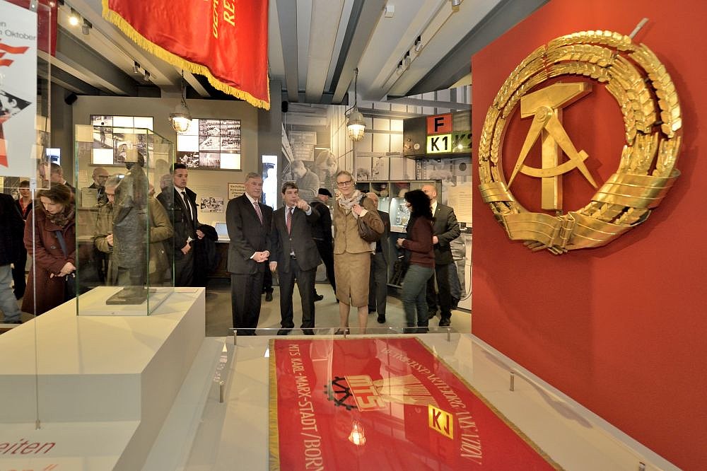Blick in die Ausstellung mit einem großen goldenen Staatswappen der DDR auf rotem Grund sowie Bundespräsident a.D. Horst Köhler und Stiftungspräsident Hütter
