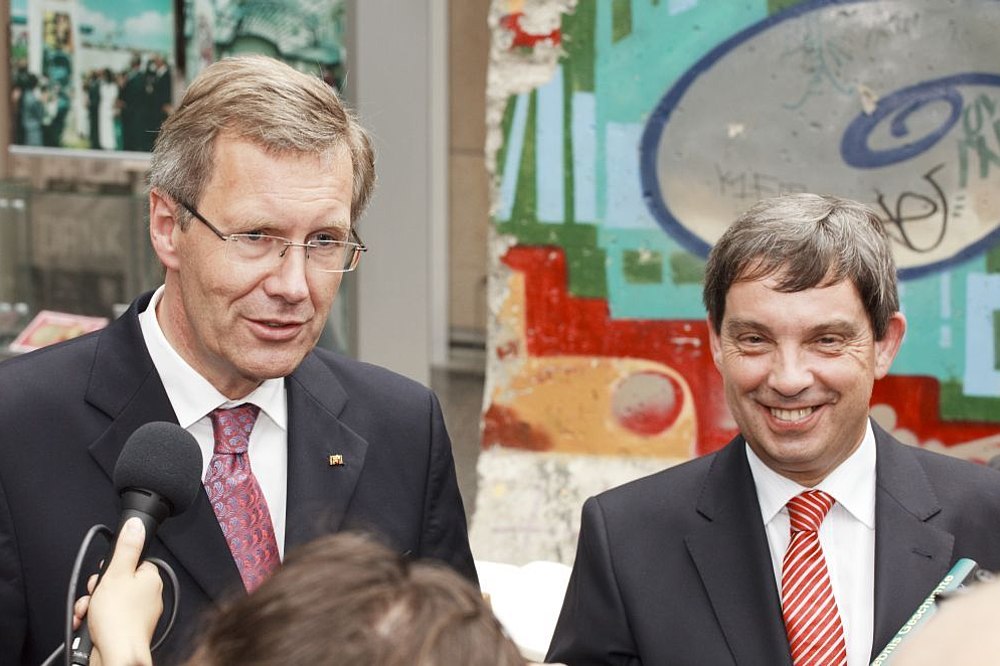 Der damalige Bundespräsident Wulff und Stiftungspräsident Hütter, im Hintergrund Bauteile der Berliner Mauer mit Graffiti, im Vordergrund Mikrofone