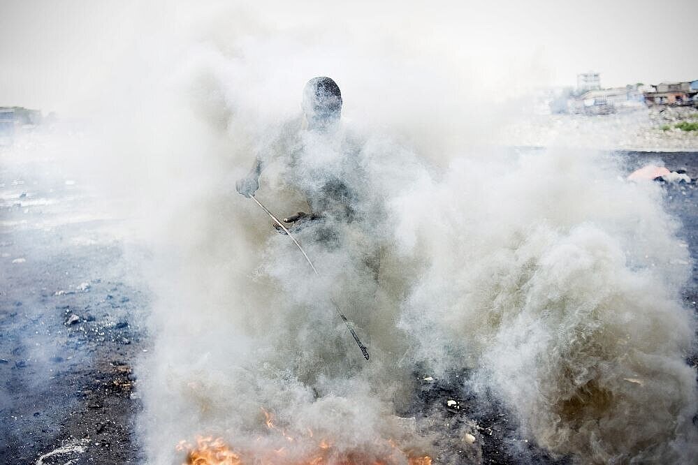 In eine dichte Rauchwolke gehüllt stochert ein Mann mit einem dünnen Stab in einem Feuer.