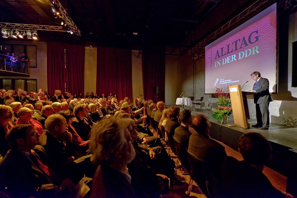Stiftungspräsident Hütter auf einer Bühne, hinter ihm das Plakat der Ausstellung Alltag in der DDR, vor der Bühne sind Stuhlreihen mit Menschen