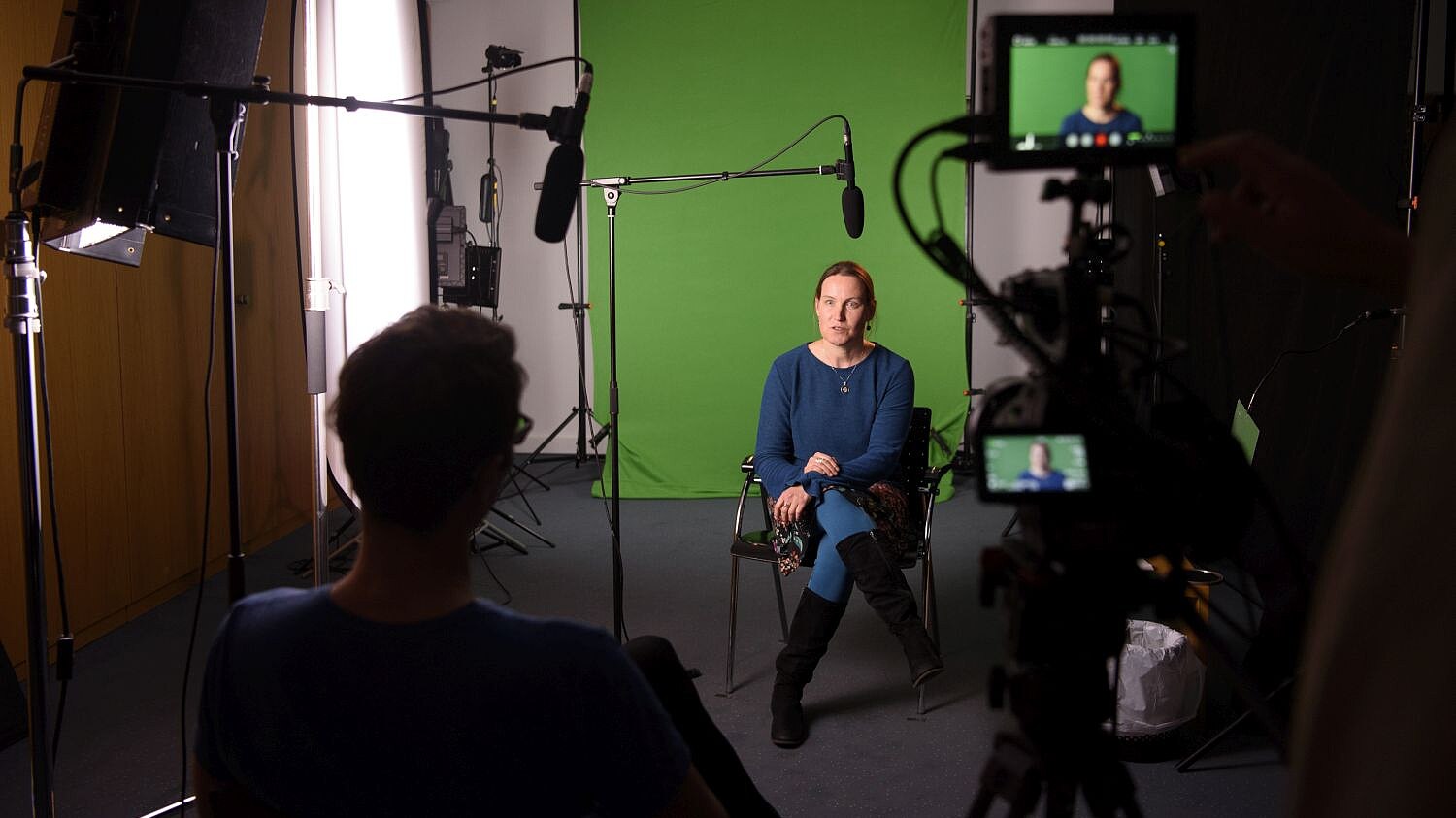 Eine mittelalte Frau sitzt auf einem Stuhl, hinter ihr ein Greenscreen, vor ihr Mikrofone, eine Videokamera, ein großer Strahler und ein Interviewer.