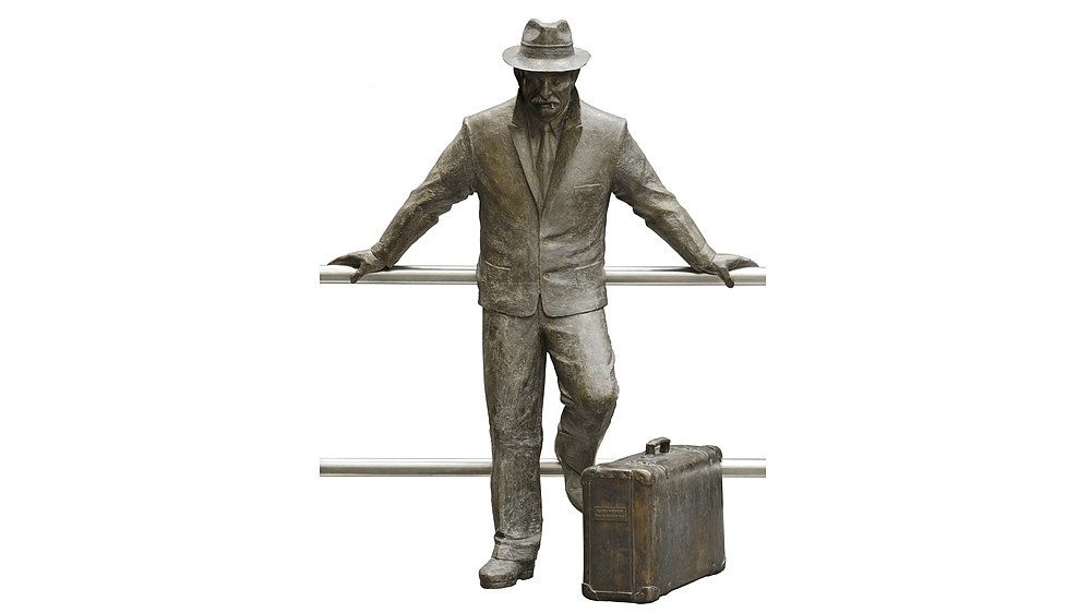 Bronzene Figur eines Mannes mit Hut, Straßenanzug, Hemd und Krawatte. In den Mundwinkeln befindet sich ein Zigarettenstummel. Das linke Bein des Mannes ist angewinkelt.