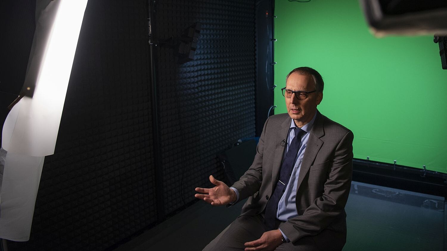 Ein älterer Mann sitzt vor einem Greenscreen. Ein Scheinwerfer ist auf ihn gerichtet, die Wand zu seiner rechten ist mit Dämm-Material verkleidet.