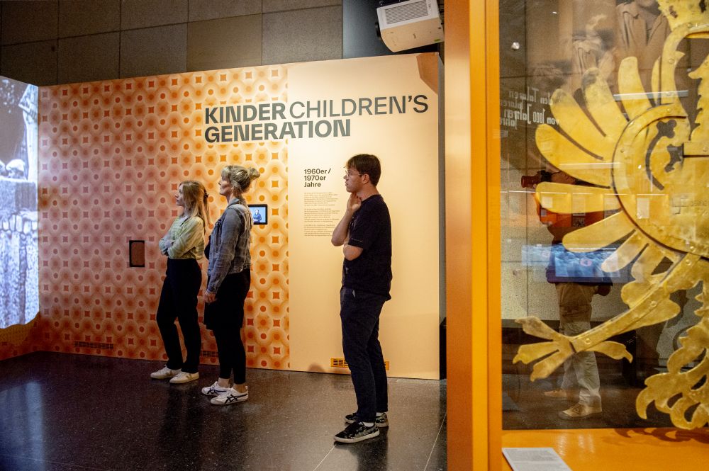 Three visitors stand in front of an orange-coloured wall and look at a screen that reads: ‘Children's generation’.