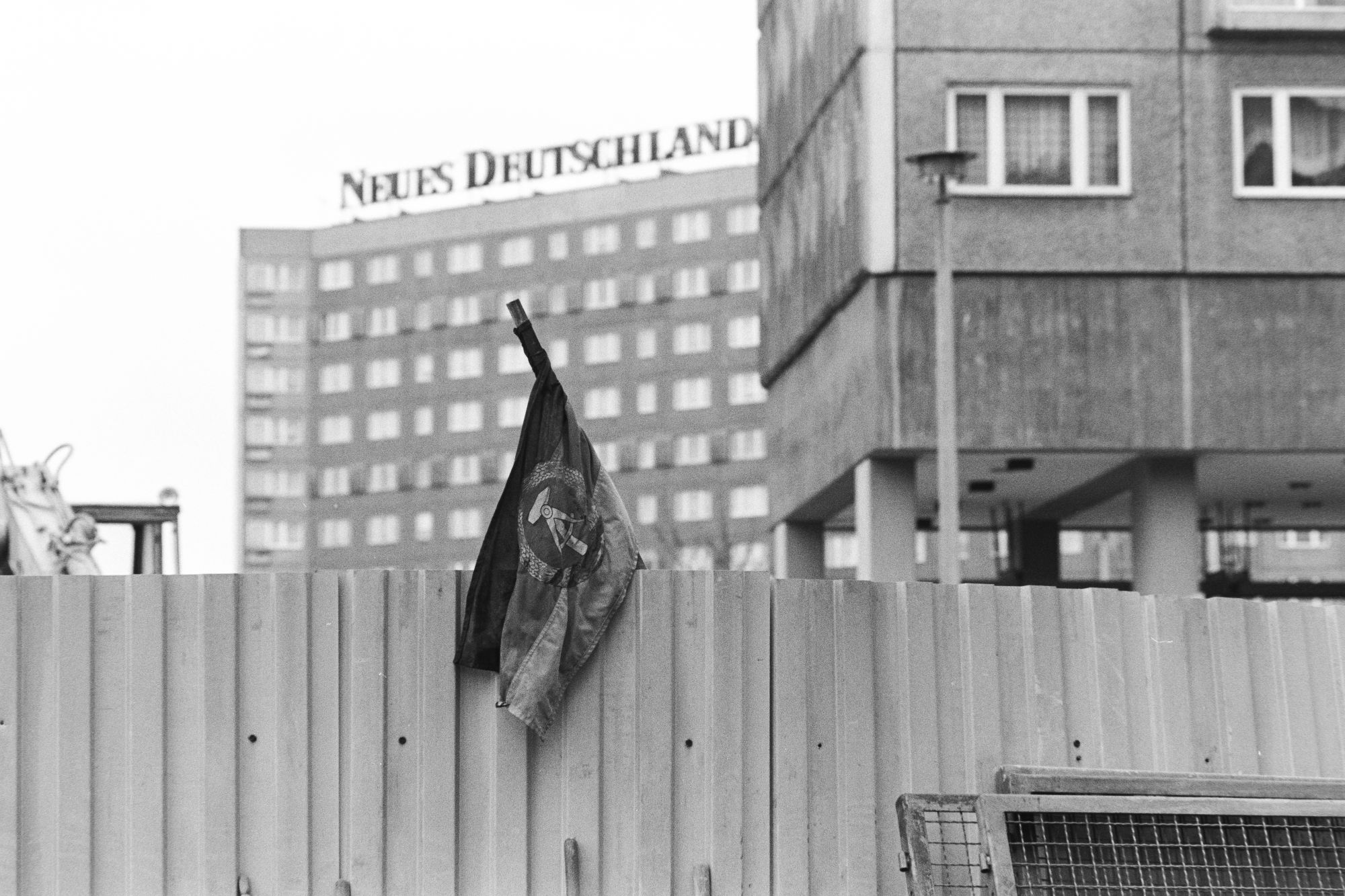 Eine DDR-Fahne über einer Mauer, dahinter ein Gebäude mit dem Schriftzug Neues Deutschland. Rechts im Bild ein Plattenbau.