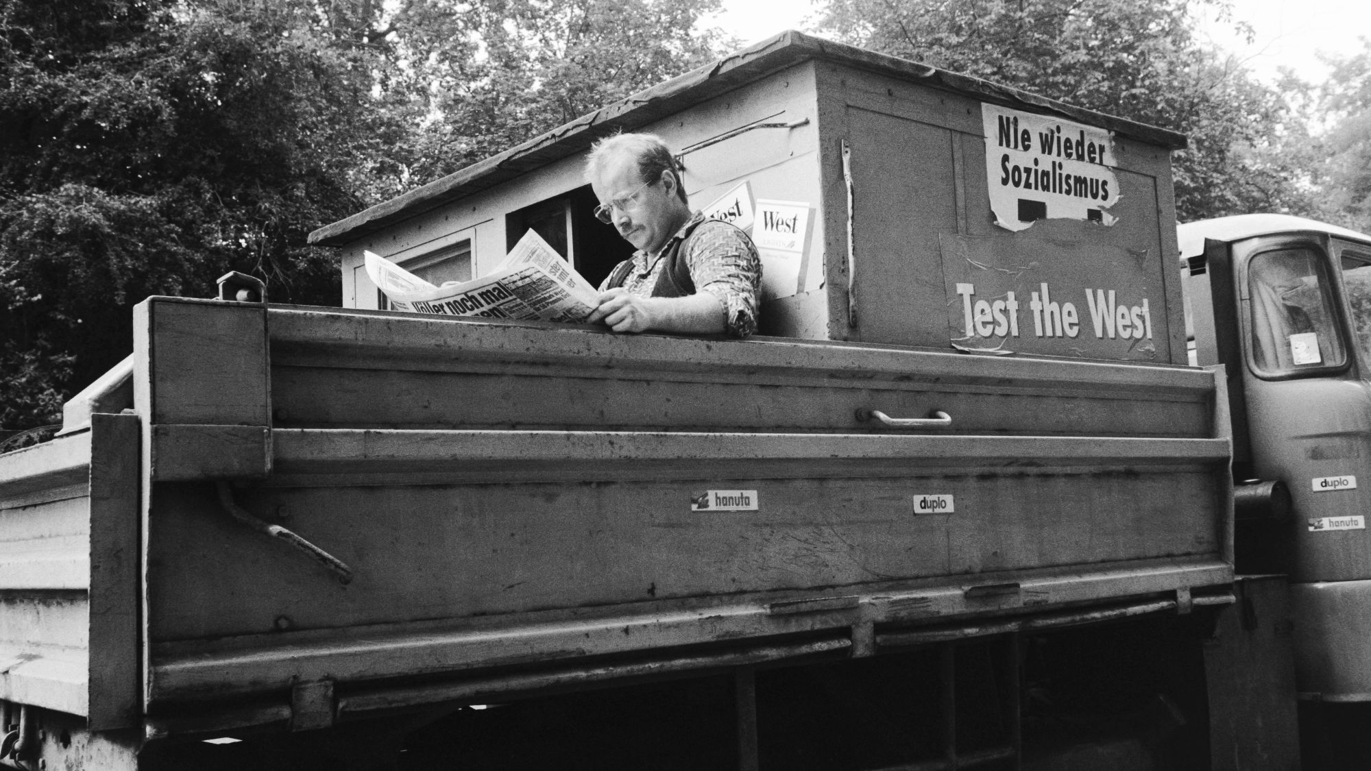 Ein Mann sitzt auf der Ladefläche eins Barkas und liest Zeitung. Auf dem Barkas Aufkleber mit Nie wieder Sozialismus und Test the West.