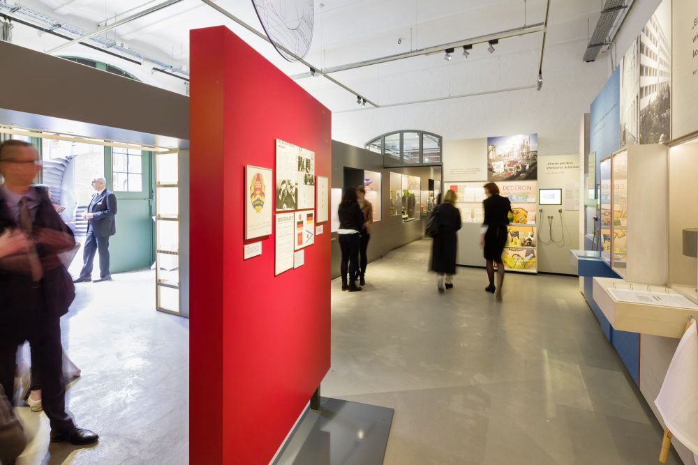 Besucher in einen großen Ausstellungsraum mit Fotos und Vitrinen und einen großen Türrahmen zu einem dahinterliegenden Raum.