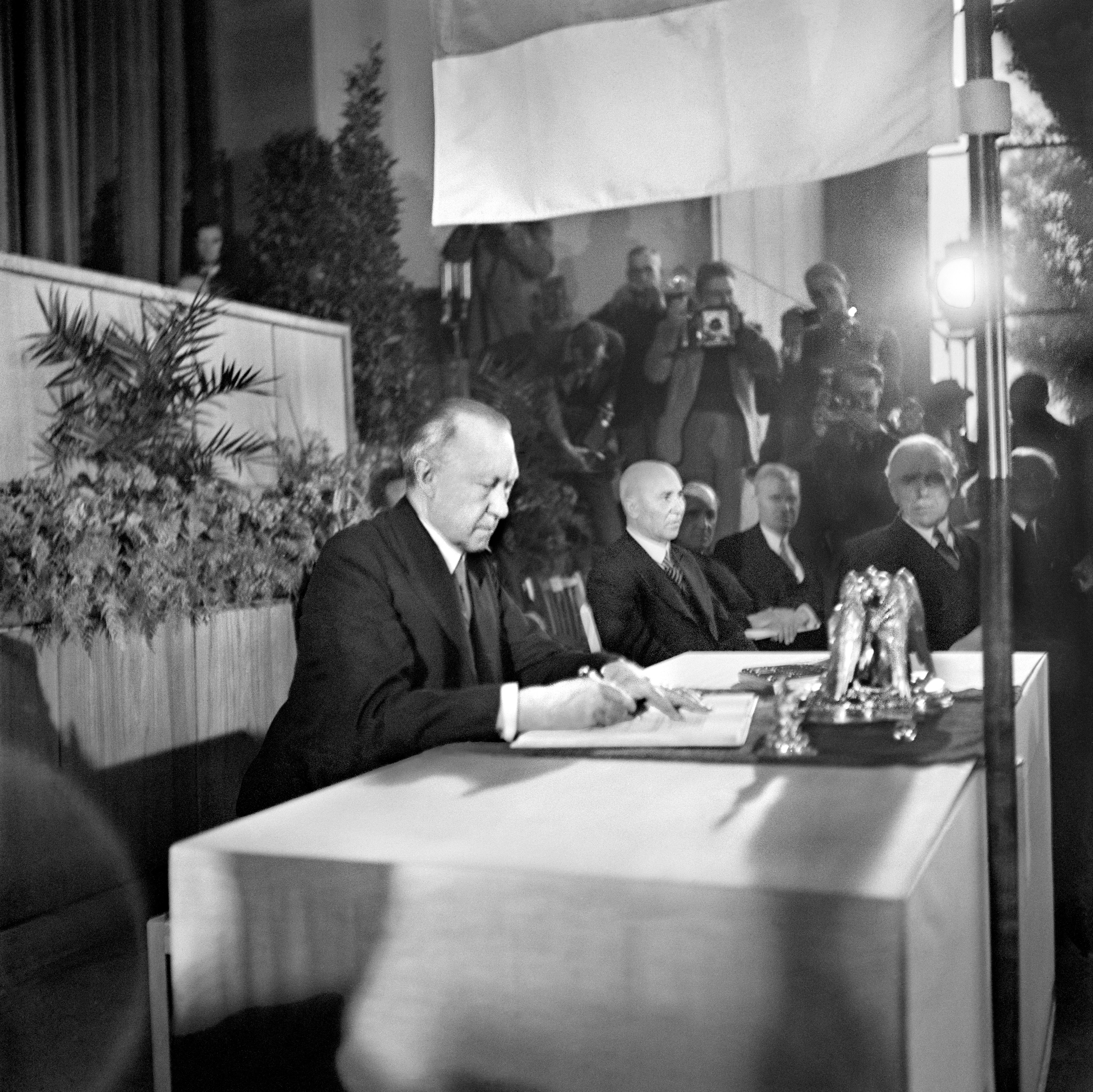 Auf dem historischen Foto sitzt Konrad Adenauer an einem Tisch und unterzeichnet das Grundgesetz. Im Hintergrund sind weitere Männer im Anzug und Fotografen zu sehen.