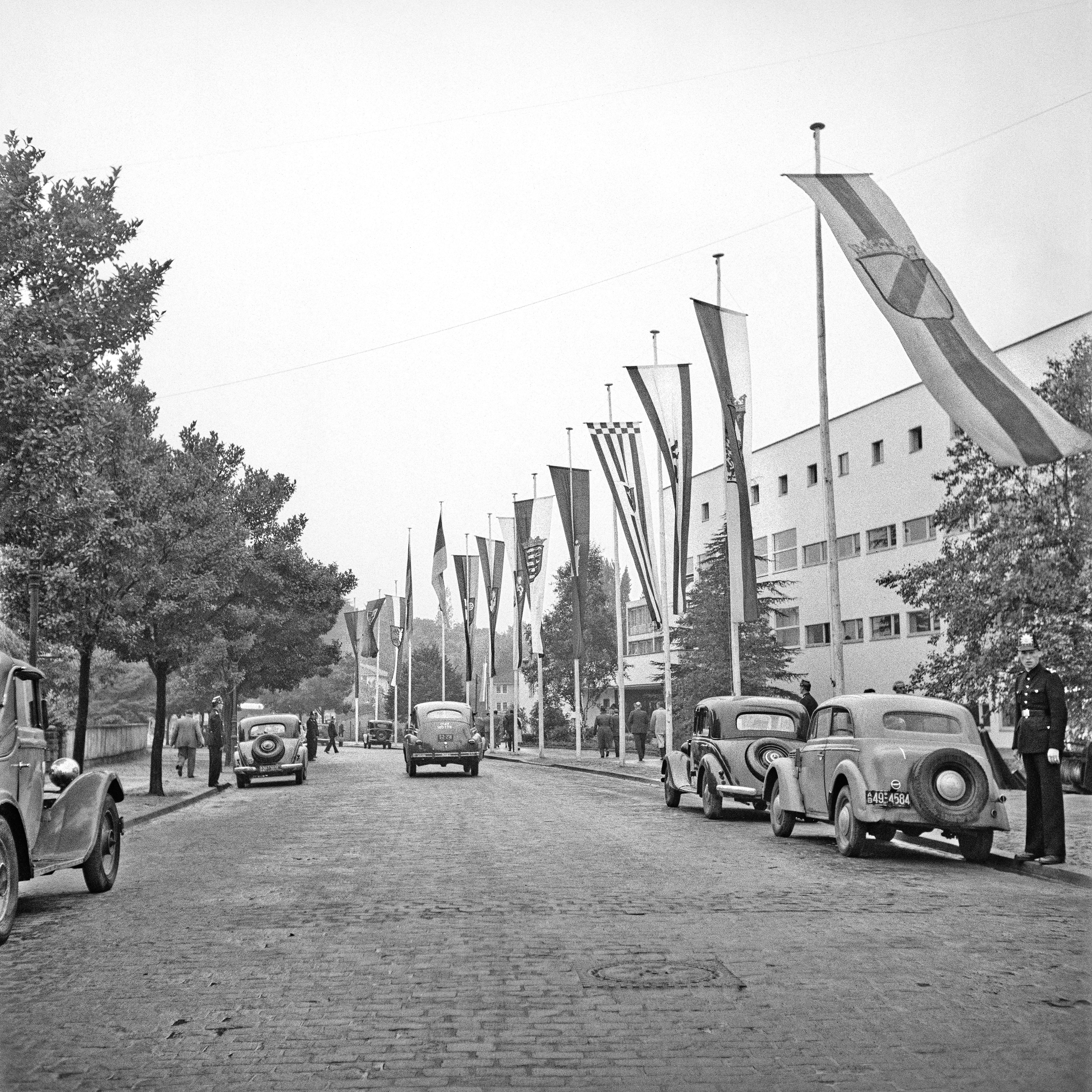 Auf dem historischen Foto ist eine Kopfsteinpflasterstraße mit mehreren Autos zu sehen. Am rechten Straßenrand und vor der Pädagogischen Akademie hängen die Fahnen der westlichen Bundesländer sowie die der Bundesrepublik an Masten.