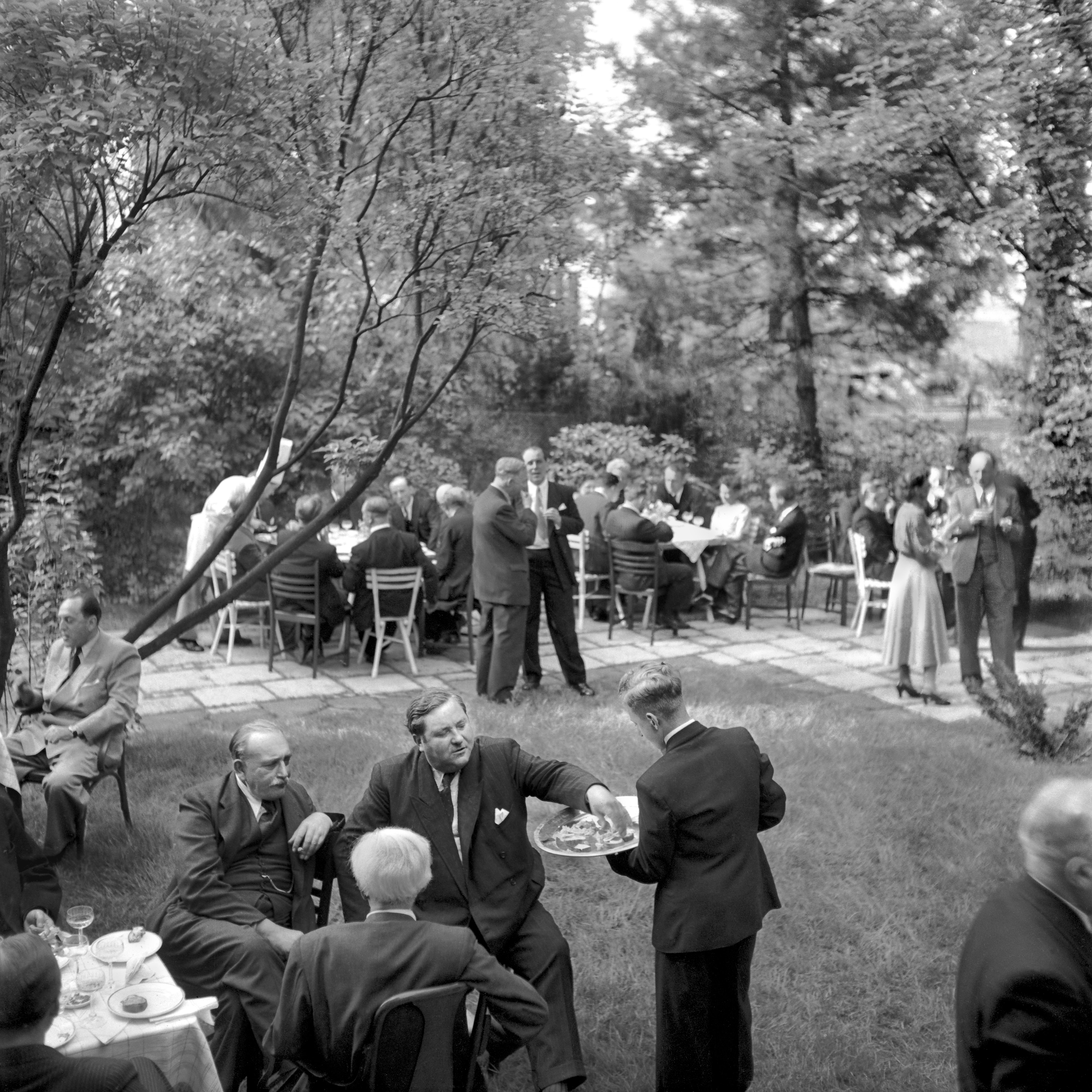 Auf dem historischen Foto greift sich Carlo Schmid Häppchen vom Tablett eines Kellners. Er sitzt zusammen mit anderen Gästen in einem Garten um gedeckte Tische herum.