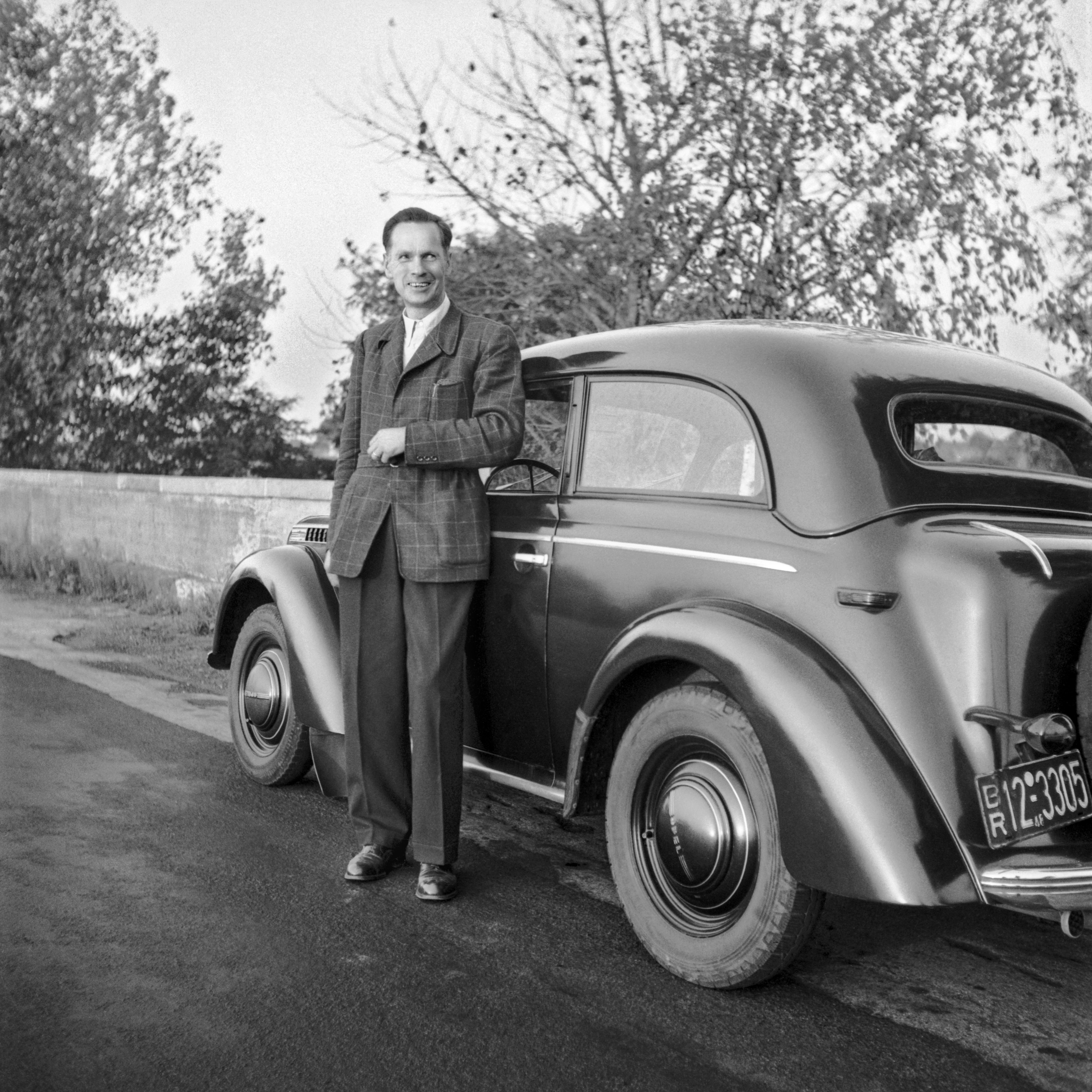 Ein junger Mann in Blazer und Stoffhose steht auf dem historischen Foto neben einem dunklen VW Käfer.