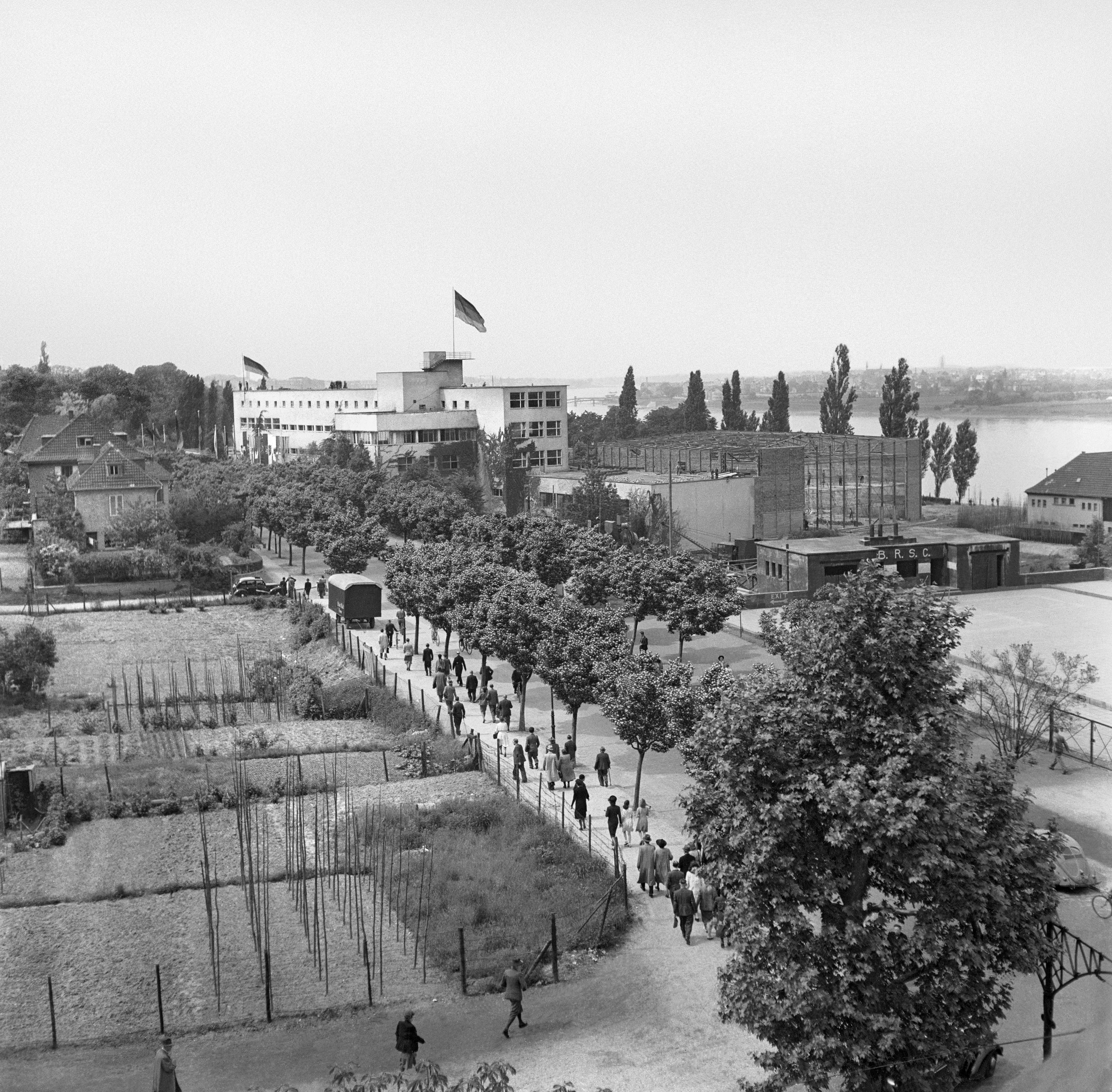 Blick von erhöhter Position auf die mit der Deutschlandfahne beflaggte Pädagogische Akademie am Bonner Rheinufer. Menschen laufen vorbei an Bäumen, Gärten und dem Rohbau des Plenarsaals des Bundestags auf die Akademie zu.