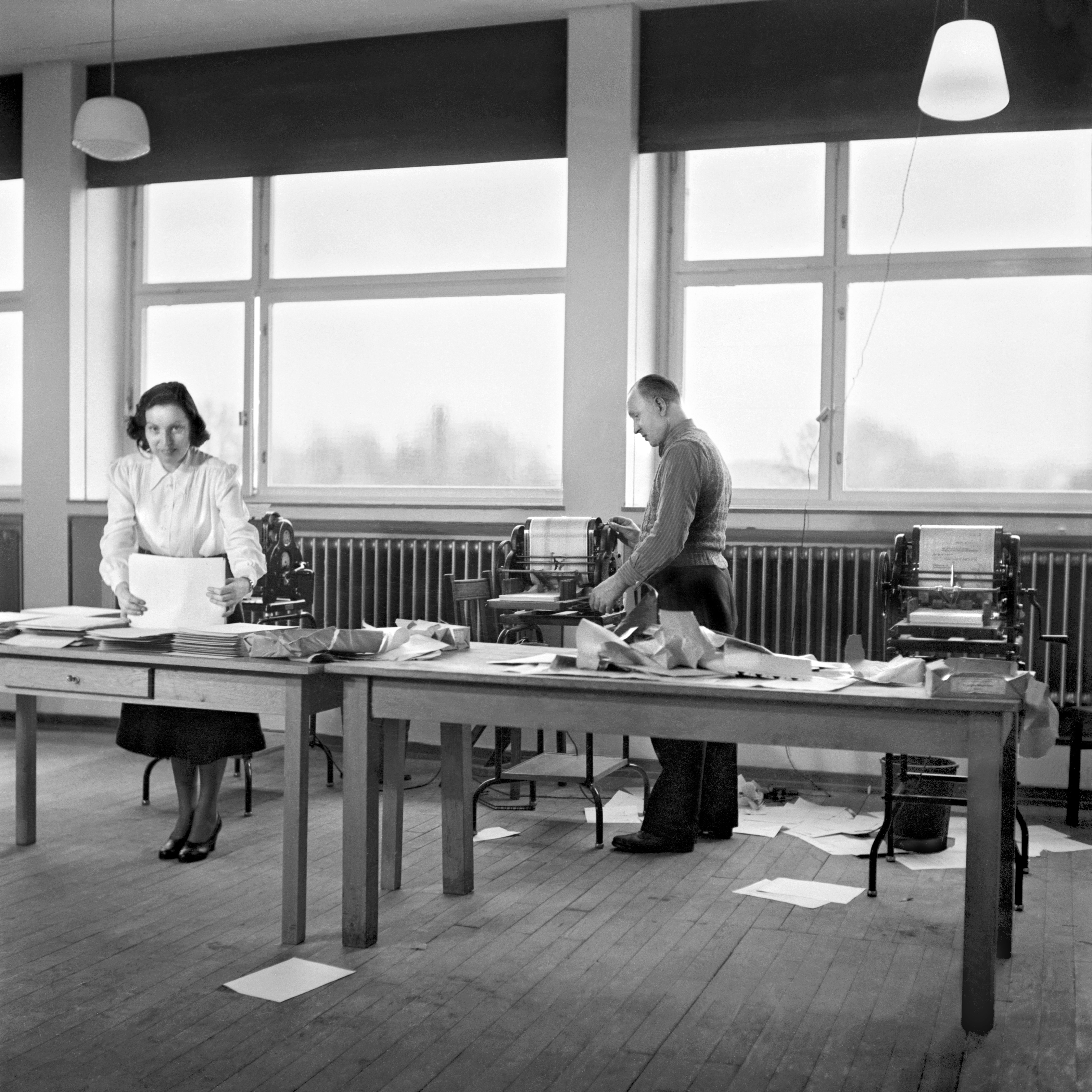 Ein Mann und eine Frau arbeiten auf dem historischen Foto an Tischen und Matrizendruckern mit den Drucksachen.