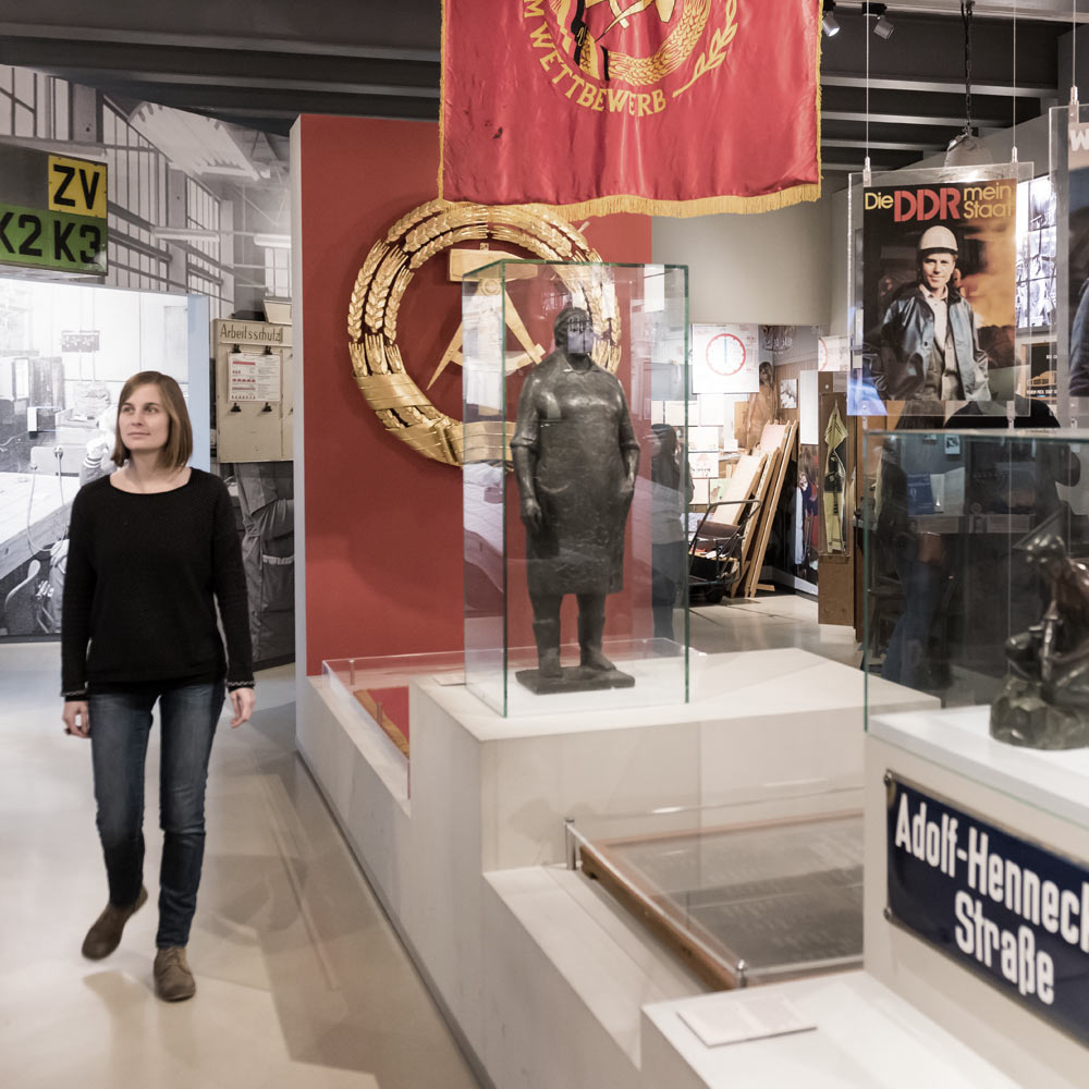  In einem Ausstellungsraum hängen Fahnen, das goldene Staatswappen der DDR auf rotem Grund und stehen Skulpturen in Glasvitrinen. Daneben geht eine junge Frau in Jeans und Pullover.