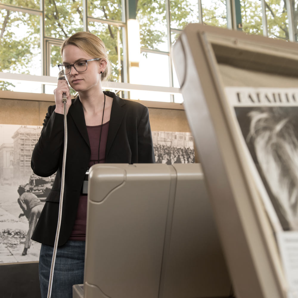 Eine junge, blode Frau in Jeans und Blazer und mit einer Brille steht mit einem Hörlöffel in der Hand zwischen der Ausstellungsarchitektur im Tränenpalast.