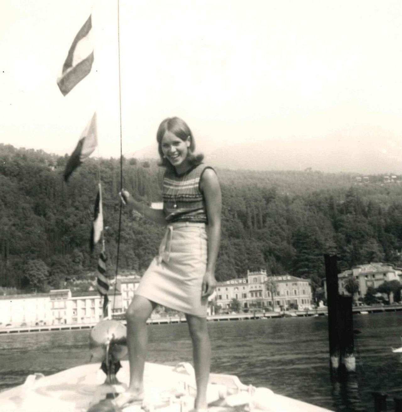 Foto, schwarz-weiß. Eine junge Frau steht am Bug eines Schiffes und lächelt in die Kamera. Sie trägt einen Rock und ein ärmelloses Oberteil. Sie hat schulterlange Haare und hält sich an einem Seil fest. Im Hintergrund sieht man Gebäude vor einem bewaldeten Hang.