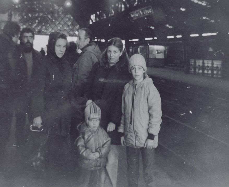 Foto, schwarz-weiß. In einem Bahnhofsgebäde stehen an einem Gleis zwei Frauen und vor ihnen stehen zwei junge Mädchen. Rechts neben ihnen sind die Gleise. Links hinter ihnen steht eine Gruppe von Männern, die sich unterhalten. 
